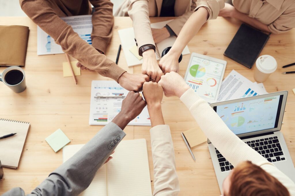 pexels photo 3184438 Top view of a diverse team fist bumping over a meeting table with paperwork and laptops, symbolizing teamwork.